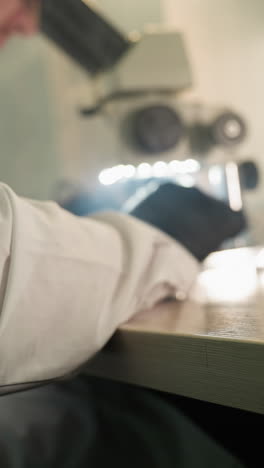 una vista trasera de un técnico en una bata de laboratorio con guantes negros usando un microscopio para inspeccionar componentes electrónicos, con otro componente visto en la mesa