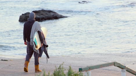 Vista-Trasera-De-Un-Hombre-Adulto-De-Mediana-Edad-Con-Una-Tabla-De-Surf-Parada-En-La-Playa-4k