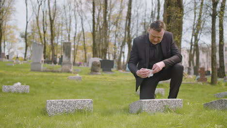 man in black suit cleaning a tombstone with a handkerchief in a graveyard