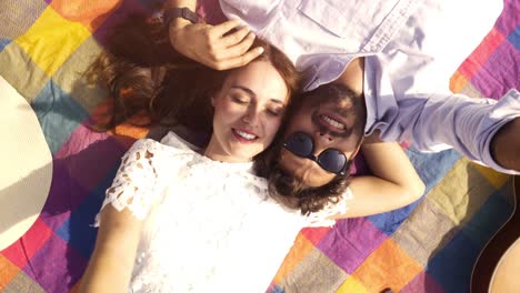 feliz y encantadora pareja joven acostada en una manta de colores en el parque mirando al cielo apuntando a las nubes romántico con gafas de sol de guitarra hermosa chica atractiva vista superior cámara giratoria en cámara lenta