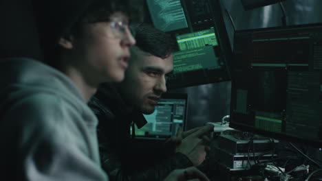 two young hackers collaborating in a dark room with multiple computer screens.