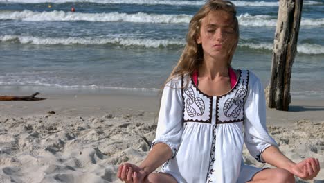 a woman wearing a white dress, sitting on the beach meditating, while beach waves crash onto the shore