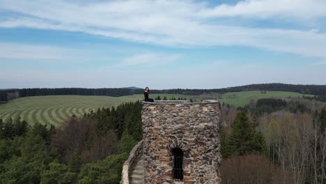 aerial view of happy young woman sitting on top of medieval tower and waving to drone camera, revealing landscape shot 50fps