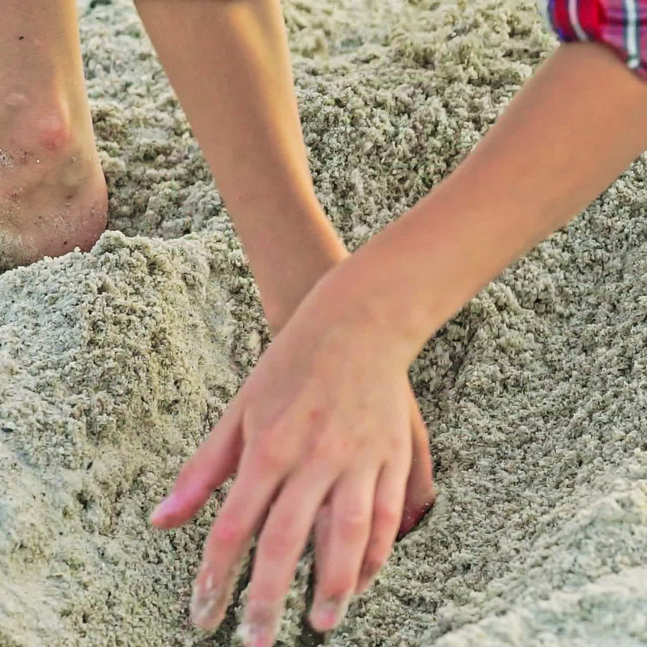 A little boy in a plaid shirt digs a bay with his hands on a sandy beach in the summer. Children's games. Close-up.