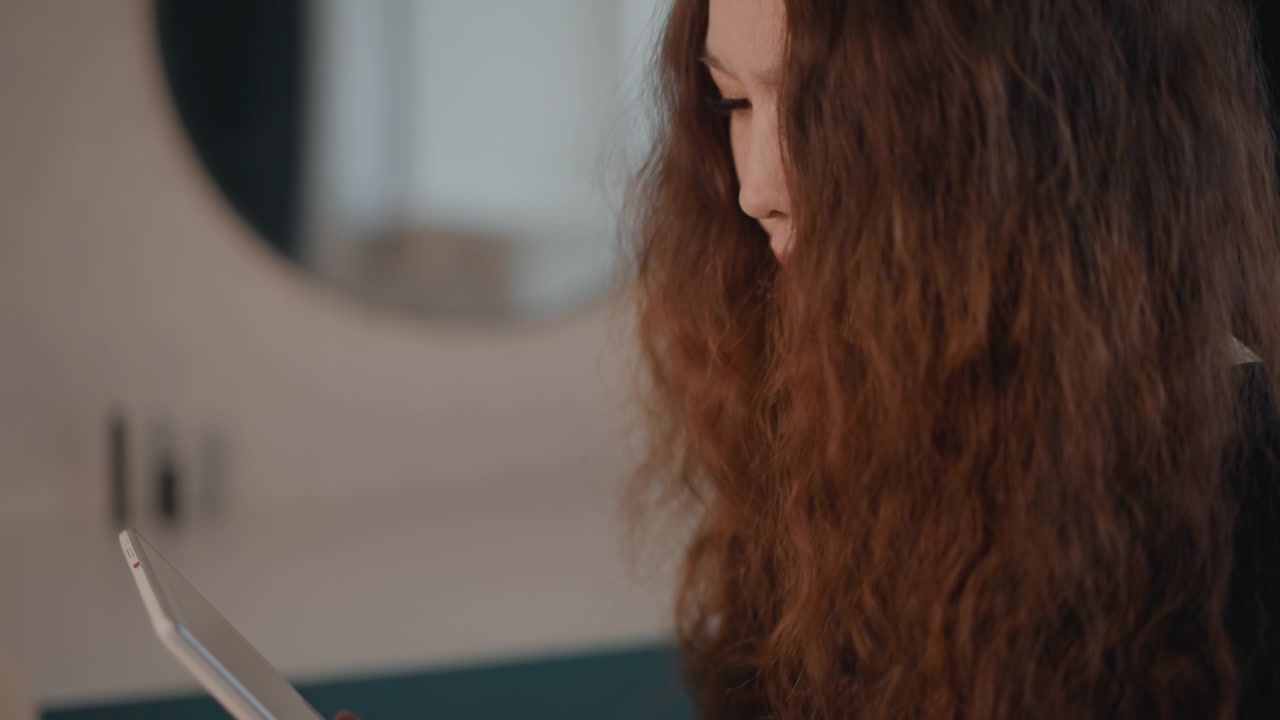 Vista de perfil de manos y parte superior del cuerpo con cabello rizado, usando una tablet y un lápiz óptico en un espacio de trabajo tranquilo, capturando movimientos deliberados de toque, escritura y dibujo con luz suave y desenfoque.