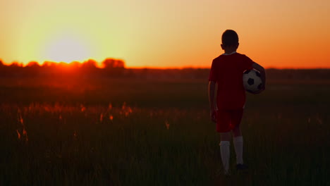 Young-football-player-goes-with-the-ball-on-the-field-dreaming-of-a-football-career-at-sunset-looking-at-the-sun.-The-camera-watches-the-boy-walking-on-the-field-at-sunset-with-a-soccer-ball.