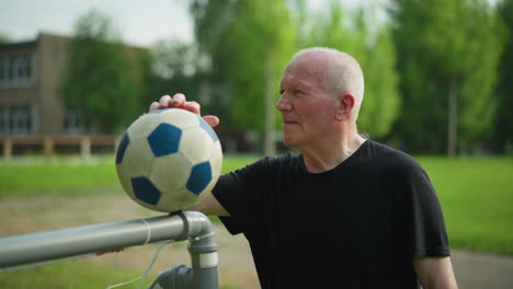 an elderly man in a black top rests his hand on a soccer ball placed on a goalpost, looking away focused, with a blurred view of a paved road and lush green grass in the background