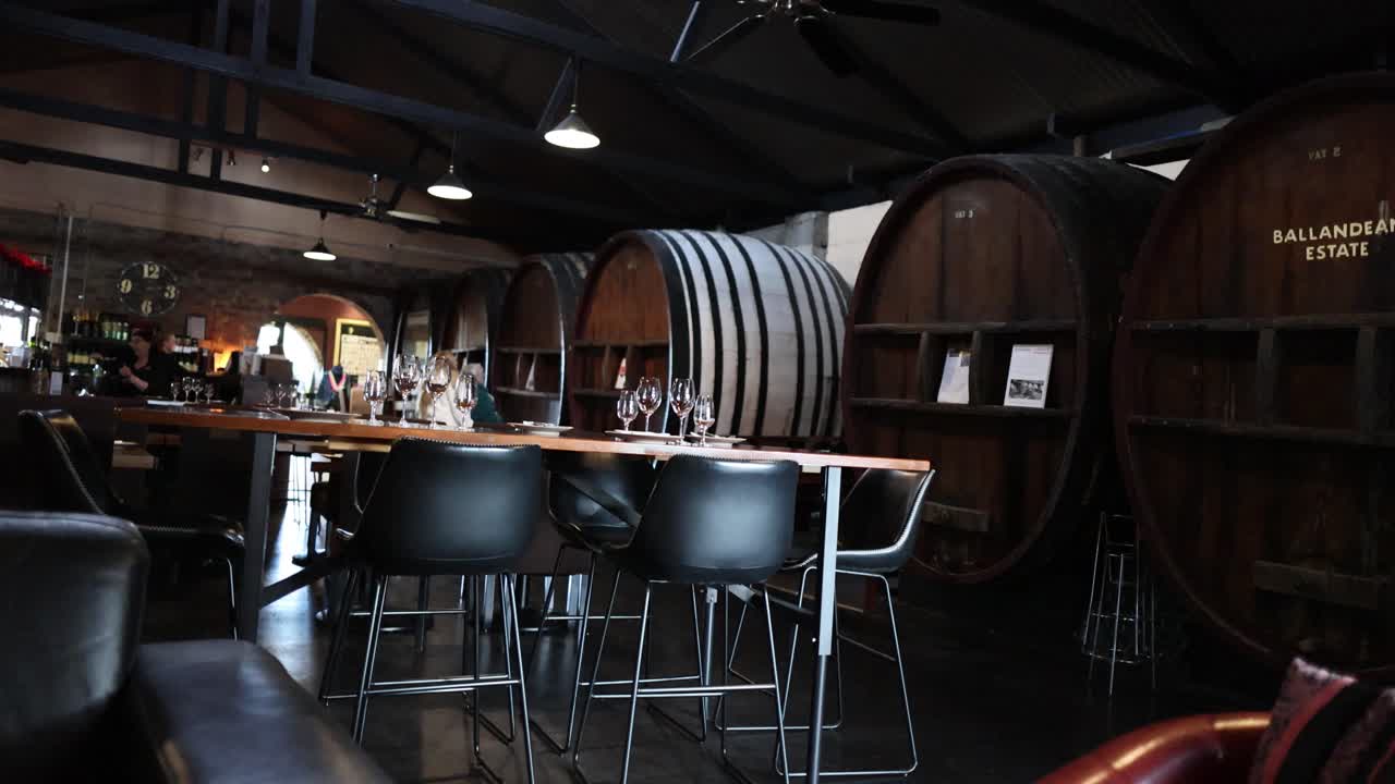 Dimly lit winery cellar with wooden barrels, tasting tables, and modern chairs, slow camera pan