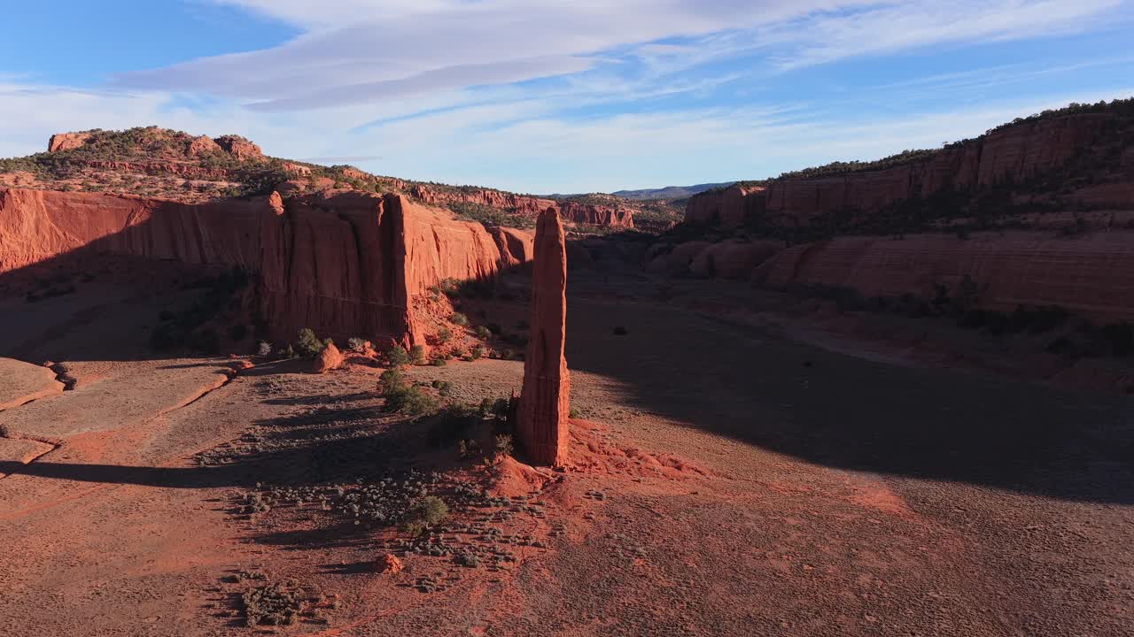 Un'orbita fluida con drone in parallasse ruota attorno a una sottile guglia di roccia rossa nel deserto, enfatizzando la sua scala verticale contro imponenti mesas sullo sfondo e un cielo blu nella remota e selvaggia zona desertica di alta quota