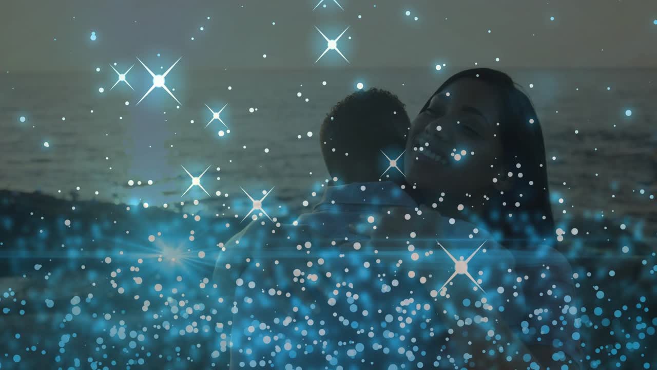Pareja abrazándose en la orilla al anochecer, la mujer inclinándose para iniciar el abrazo, con un bokeh azul flotando sobre ellos para anuncios.