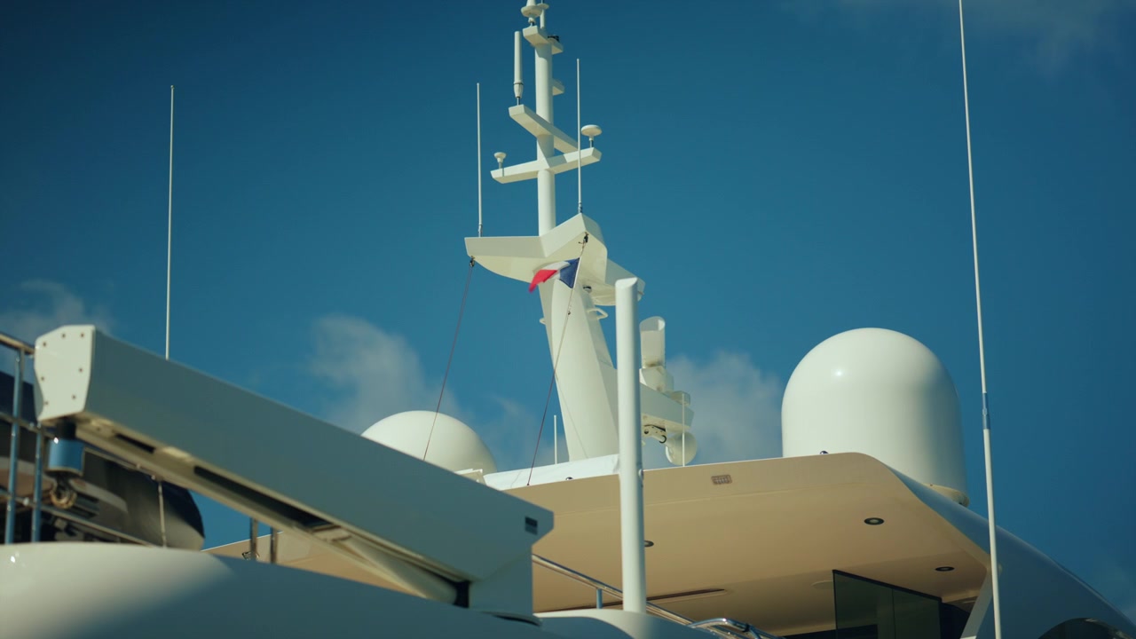 Top section of a luxury yacht showing its mast with radars, antennas, and a french flag against a blue sky