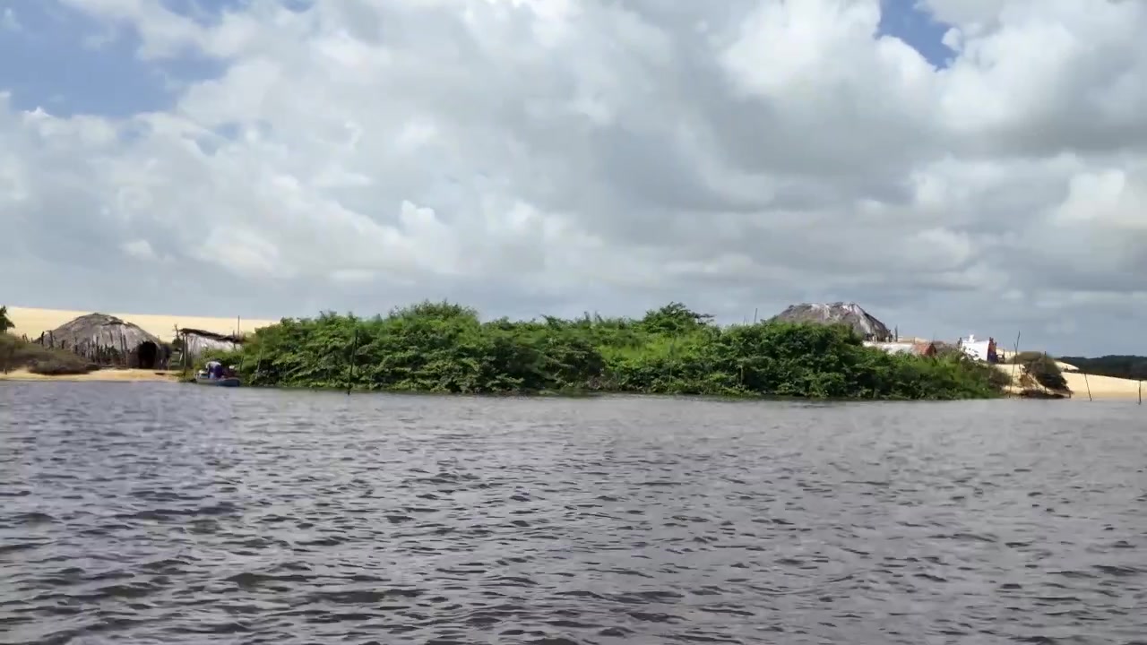Lavvinklet vannbilde som viser rustikke hytter, grønn vegetasjon og hvite sanddyner ved kanten av Preguiça-elven under en skyet himmel i Maranhão, Brasil.