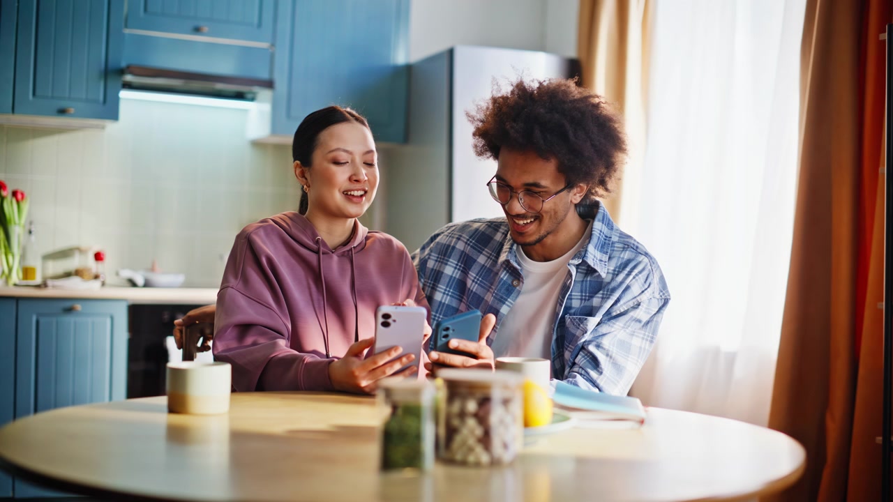 Young couple comparing smartphones at home kitchen. Cheerful pair laughing