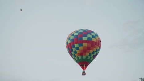 two colorful checkered hot air balloons carrying passengers ascending slowly with visible burner flames over lush green field bordered by forest and parked trailer vehicle under pastel dusk sky