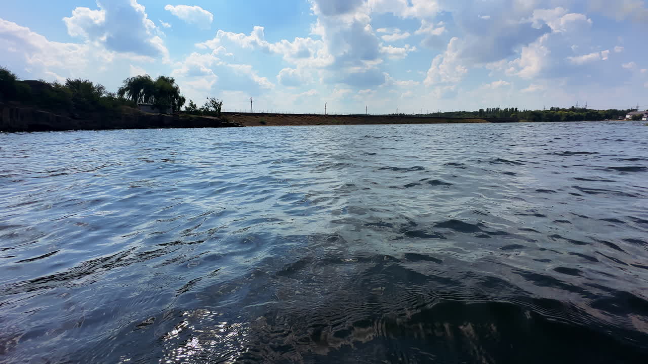 Surface of river at sunny daytime. Waving waters close up. Bank and cloudy sky at backdrop.