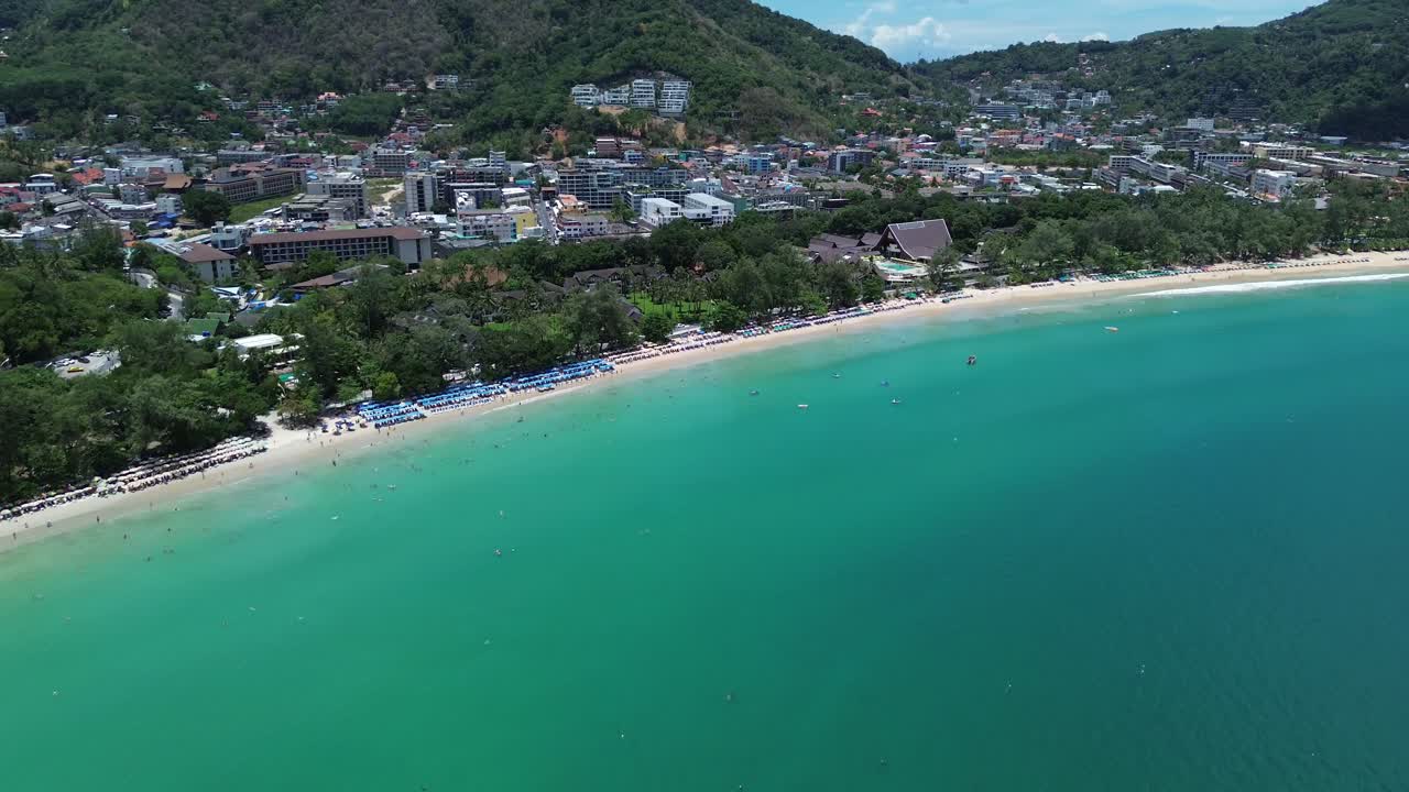 Aerial zoom in over emerald waters toward beach, trees and mountain cityscape under blue sky in Phuket