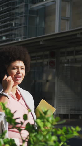 plus size biracial woman talking on smartphone and holding notebooks in city