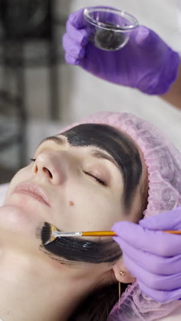 a doctor-cosmetologist applies a black mask with a brush to cleanse and moisturize the skin of the face in the beauty center. close-up. vertical video