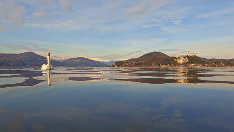 dolly fuera ángulo bajo vista de la superficie del agua de un maravilloso cisne nadando en el agua del lago con una fortaleza en el fondo y montañas nevadas