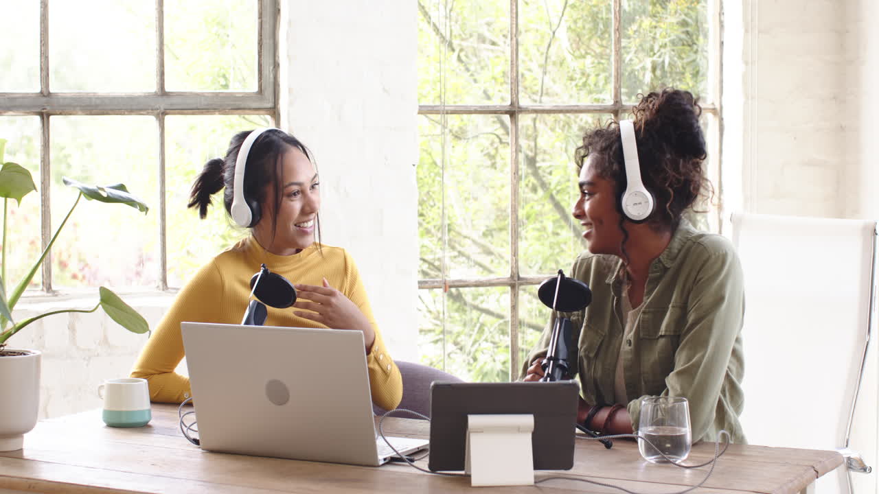 Two multiracial female colleagues recording podcast at home, with headphones and using microphones