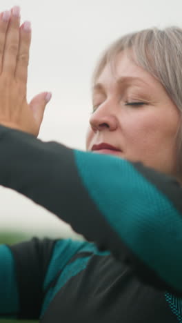 primer plano de una mujer de mediana edad con los brazos levantados, las manos unidas en una postura meditativa, practicando la atención plena al aire libre en un campo de hierba brumosa bajo un cielo nublado