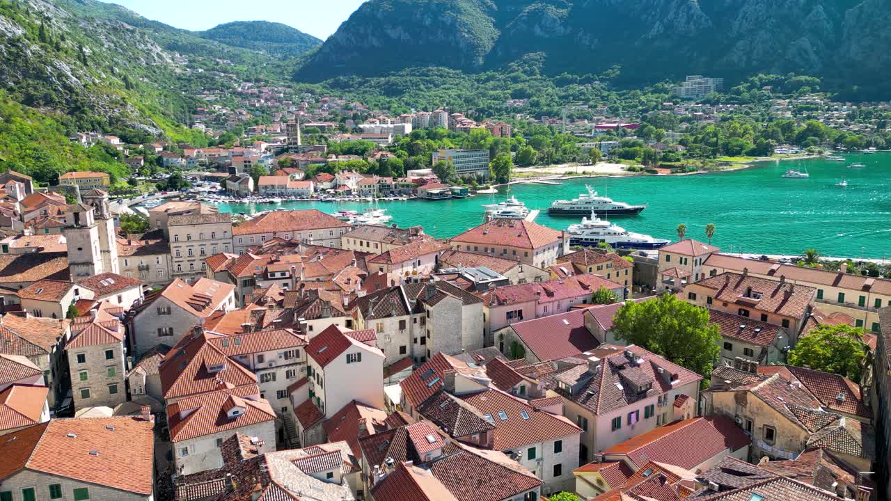 Drone passing forward above city center of Kotor. Famous travel destination in Montenegro. Luxury yachts and boats. Turquoise water.