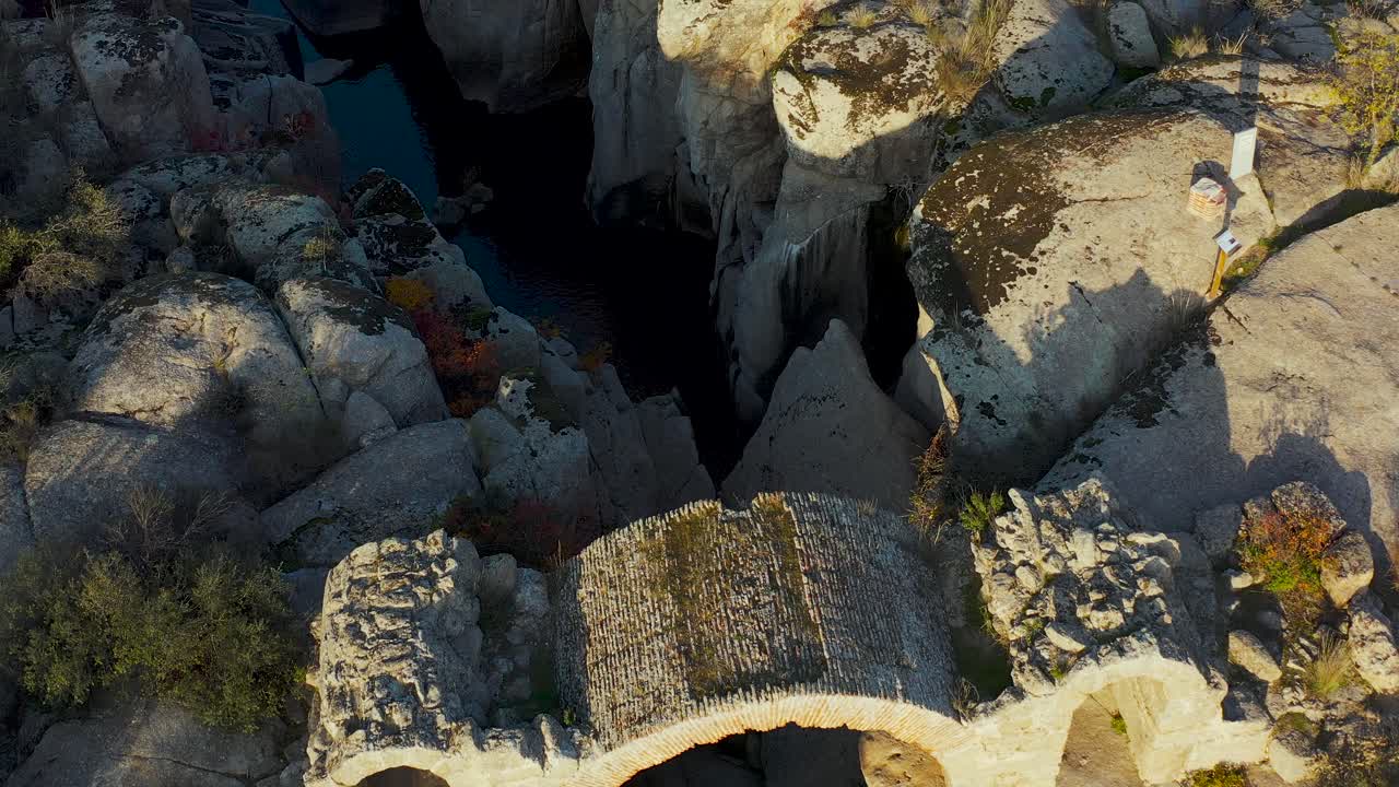 Drone frontal and overhead view at golden hour reveals the ruins of an ancient stone bridge with three arches over a rocky river, surrounded by nature and historic charm in warm light
