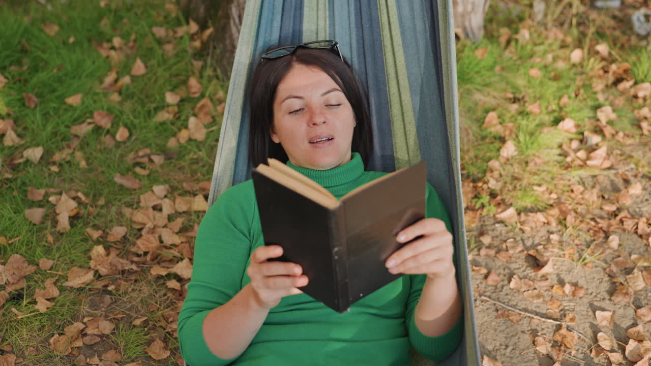 Vrouw leest buiten. Persoon ligt vredig in hangmat omringd door kleurrijk gebladerte. Vrouw verdiept zich in leesstof in de open lucht te midden van een levendige herfstachtige omgeving en bomen
