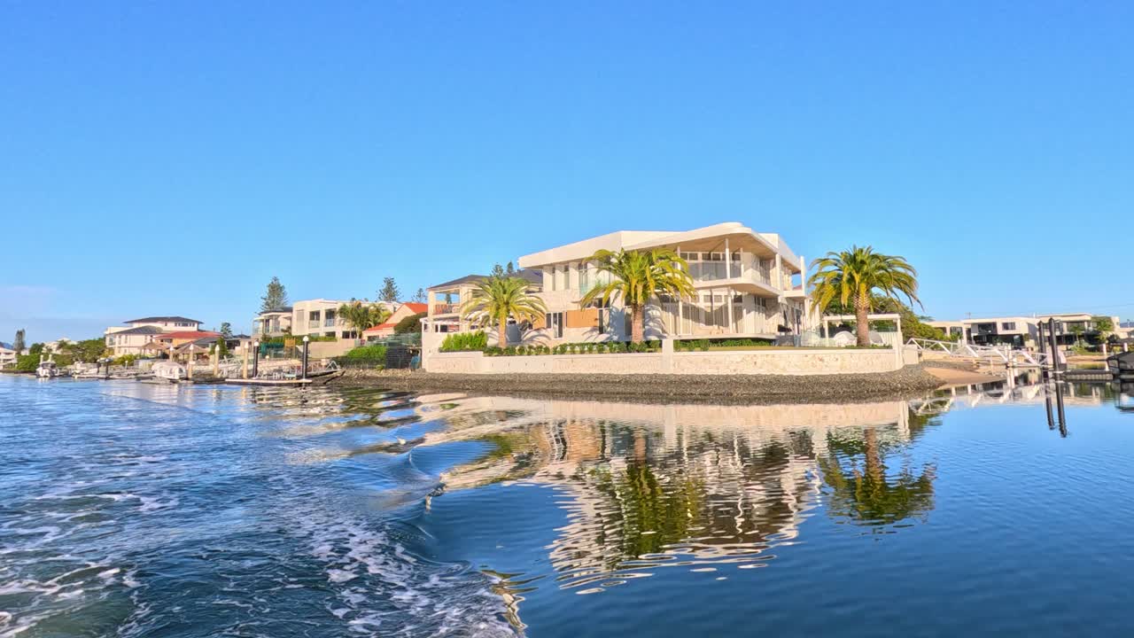 A camera glides smoothly over calm river water, revealing a modern luxury house with palm trees under bright daylight in a Gold Coast suburb