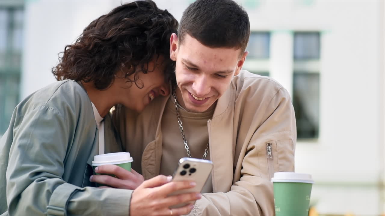 A happy couple outdoors near a cafe. Looking in the phone, laughing, talking, coffee. Autumn atmosphere. Slow motion