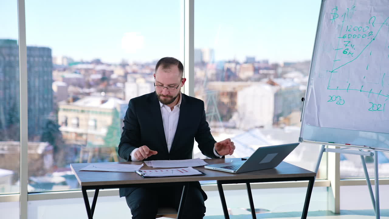 Portrait of a businessman at workplace. Bearded office worker in suit sitting at the table with a laptop on window background in the city.