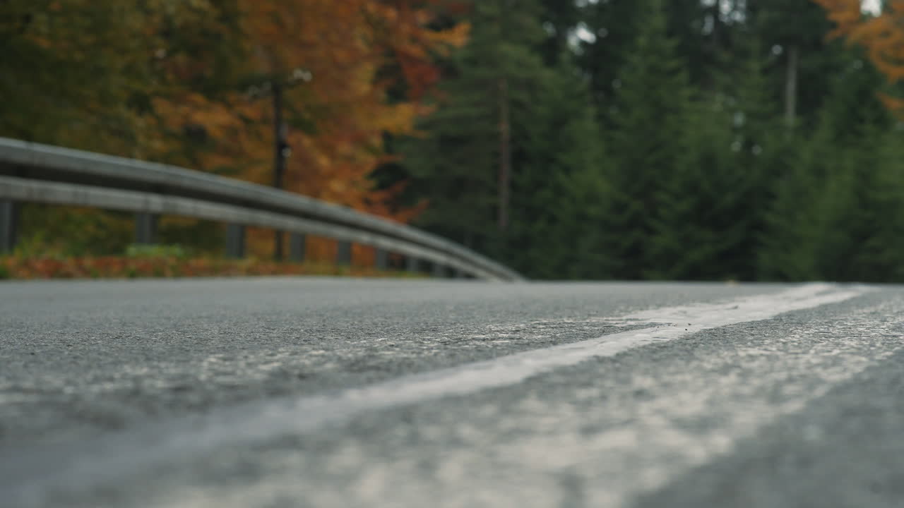 Motorcyclist riding through frame on scenic forest road in autumn. Low angle static shot. Asphalt in the foreground with focus pull. Right to left.