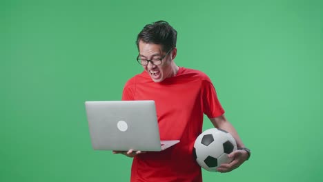 un hombre asiático con una pelota está gritando gol alegrándose por el equipo ganador mientras anima el fútbol en una computadora portátil en el estudio de pantalla verde