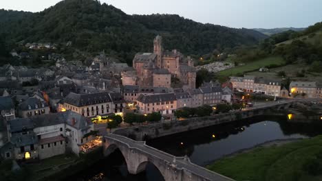 drone aerial view in france countryside small old medieval town and a castle over stone bridge on a river in a green mountain valley forest at sunset in estaing