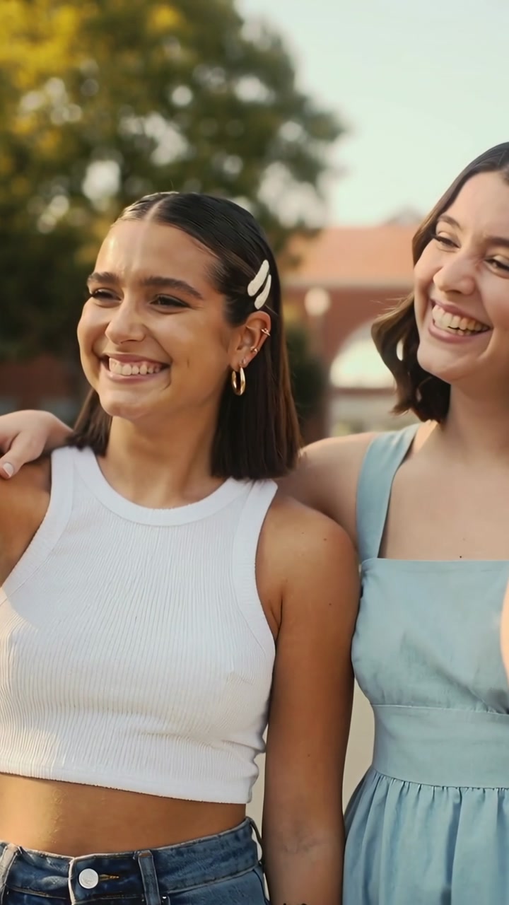 Vertical video: Walking two women laughing, left arm out for selfie on campus in tank top and dress