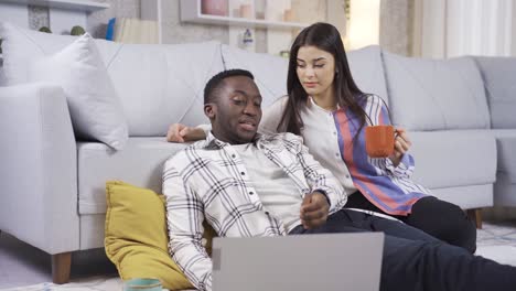 un joven africano trabajando desde una computadora portátil, charlando con su esposa blanca, diciendo algo.