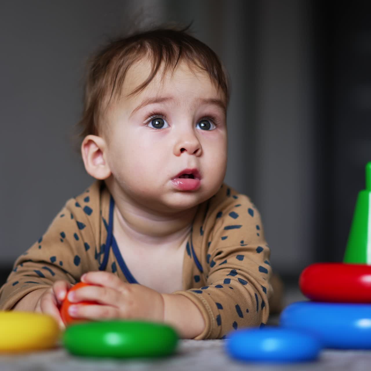 Adorable baby boy lies on the floor looking up with surprise. Child touches the parts of toy pyramid lying unfold in front of him