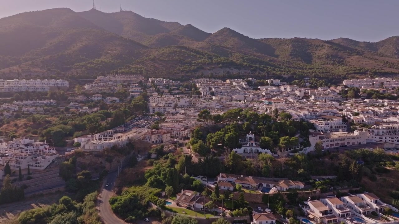 Riprese drone che si alzano su Benalmádena Pueblo, rivelando fitti tetti imbiancati, colline boscose, una cappella in cima a una collina e lo sfondo della Sierra che brilla sotto la foschia estiva