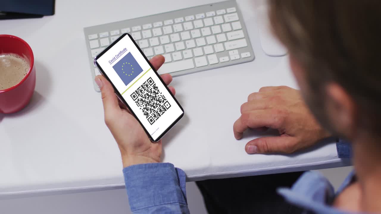 Man at desk holding smartphone with covid vaccination certificate, eu flag and qr code on screen