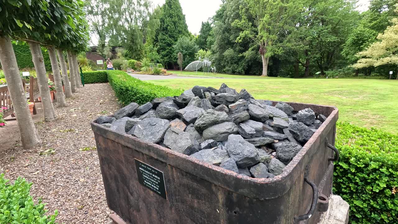 A stationary wooden coal cart filled with coal sits outdoors in a lush botanical garden, captured in daylight with slow lateral camera movement