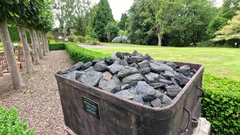 a stationary wooden coal cart filled with coal sits outdoors in a lush botanical garden, captured in daylight with slow lateral camera movement