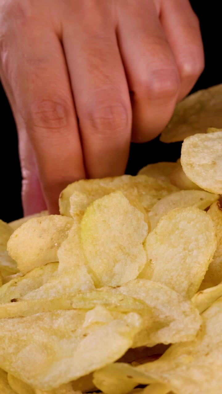 Mão humana seleciona batata frita de uma pilha, close-up, bem iluminado, fundo preto, câmera suave