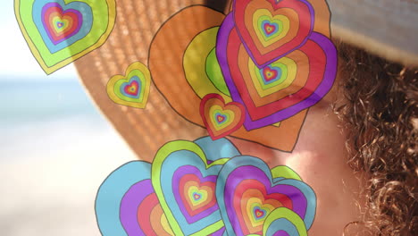 animation of colourful hearts over biracial woman wearing hat on beach