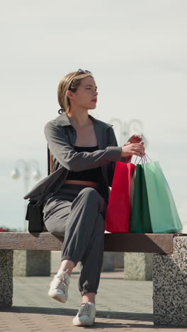 dama con tres bolsas de compras camina hacia bancos al aire libre, coloca cuidadosamente sus bolsas en el banco, se sienta, ajusta su bolso de mano negro, y cruza la pierna, mientras cruza su pierna
