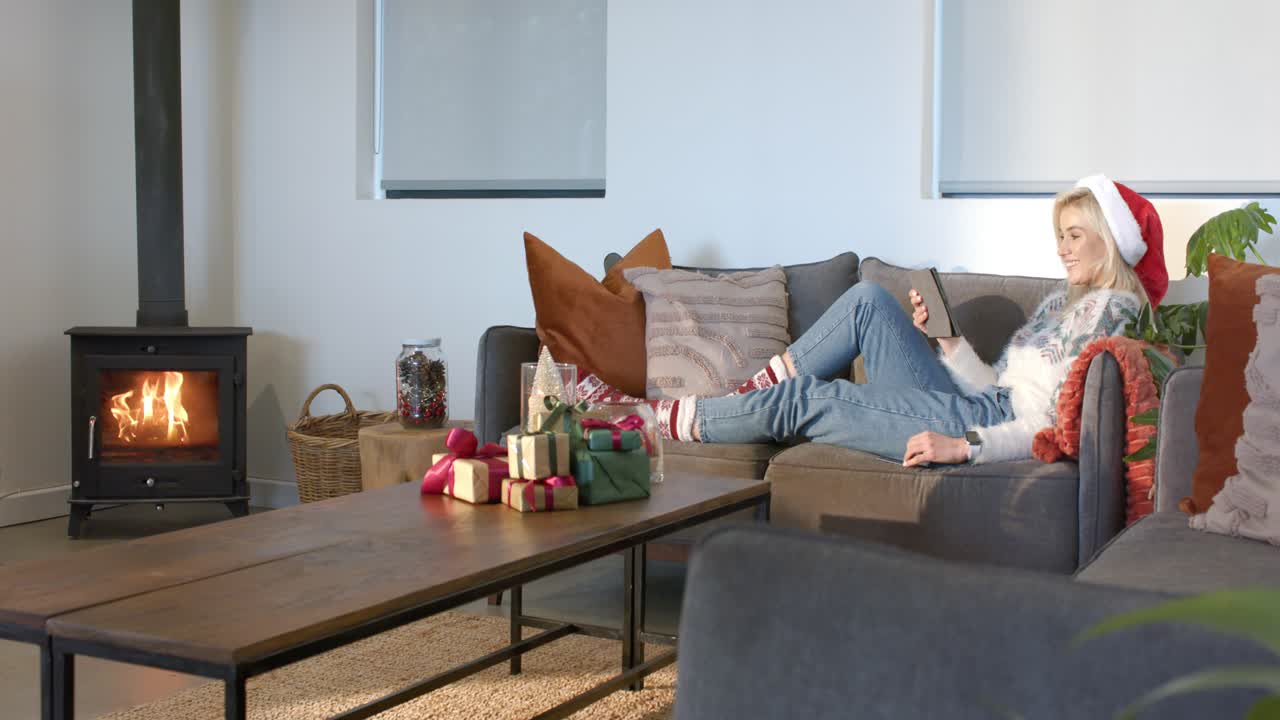 Eine Frau im Weihnachtspullover entspannt auf dem Sofa neben dem Ofen, scrollt auf ihrem Handy, greift nach dem Tablet, mit Platz für Text.