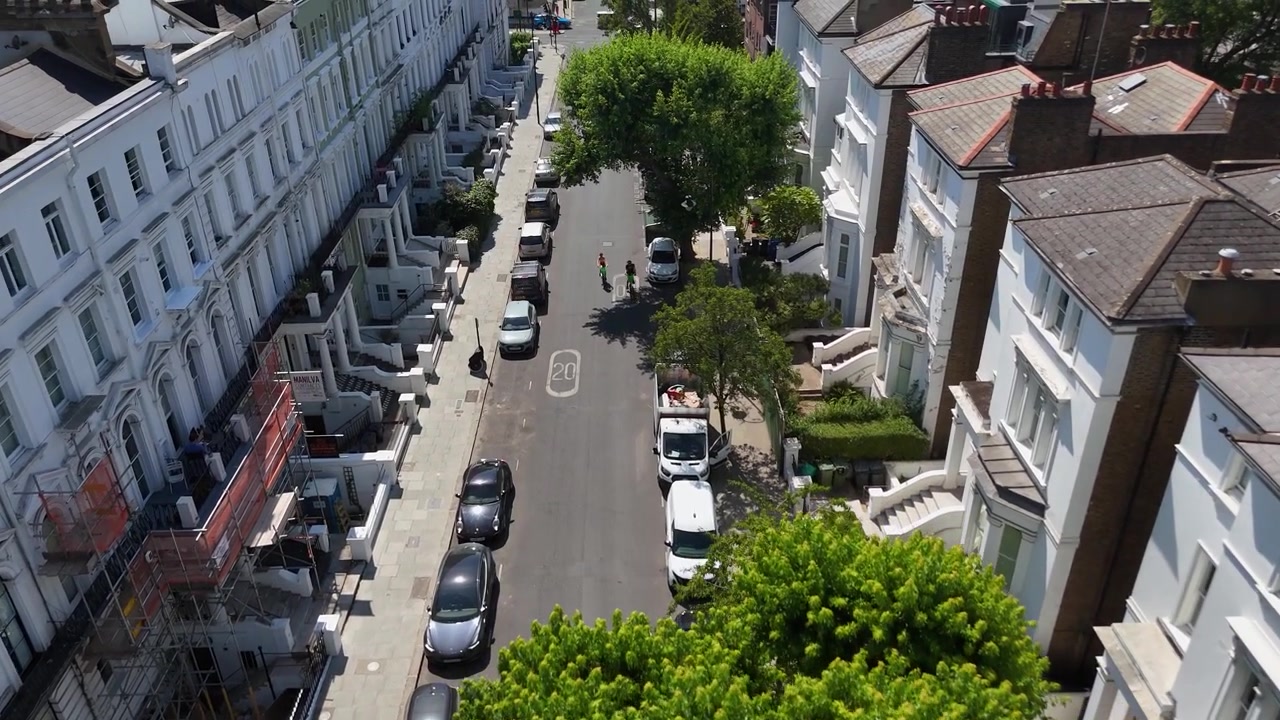 Strada residenziale alberata che si snoda tra file di case in stucco bianco con balconi e ampliamenti sul tetto