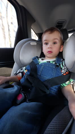 calm two-year-old toddler in jeans romper sits in baby car seat. lovely boy travelling comfortably and safe. low angle view. vertical video.