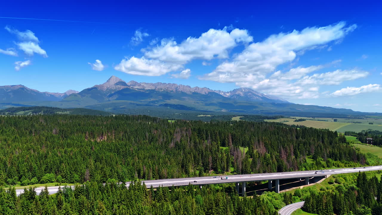 Flying closer to a freeway in the beautiful nature. Pine tree woods surround the highway. Tatra mountains at backdrop. Slovakia