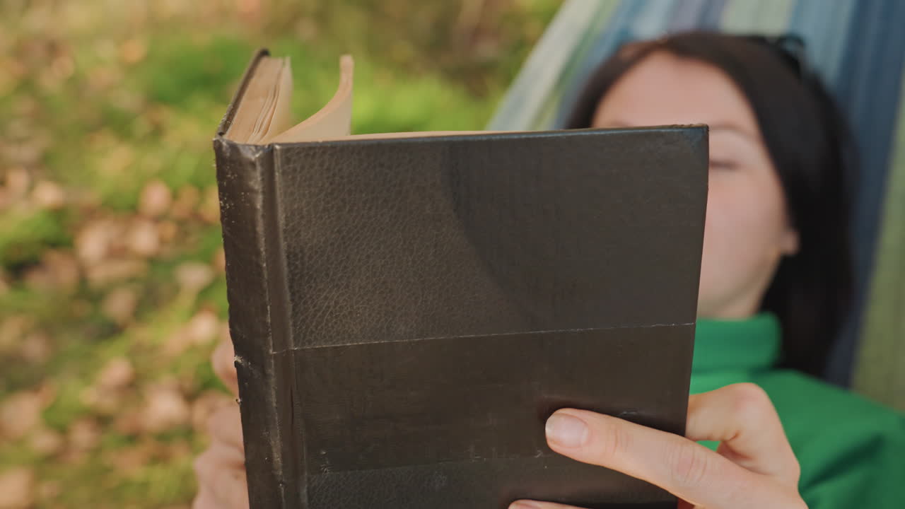 Donna in amaca che tiene un libro nero, primo piano su mani e copertina, pelle testurizzata, foglie autunnali sotto i piedi, gira le pagine con attenzione, espressione pensierosa, prospettiva neutra per scrittore o viaggiatore.