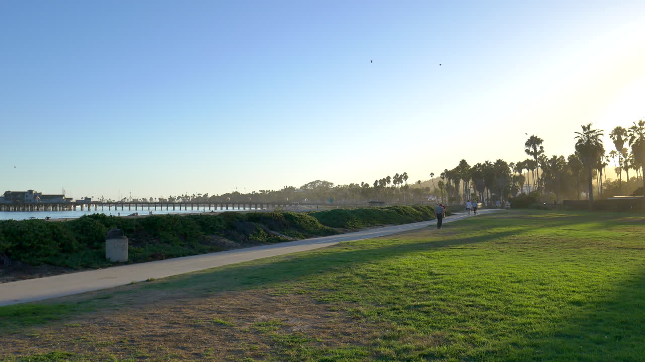 mirando alrededor de la playa con la gente caminando a lo largo de los senderos para bicicletas y palmeras en silueta contra una brillante puesta de sol en la hermosa ciudad de santa bárbara, california
